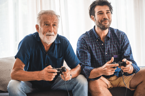 Elderly father and middle aged son play playstation together this Father's Day